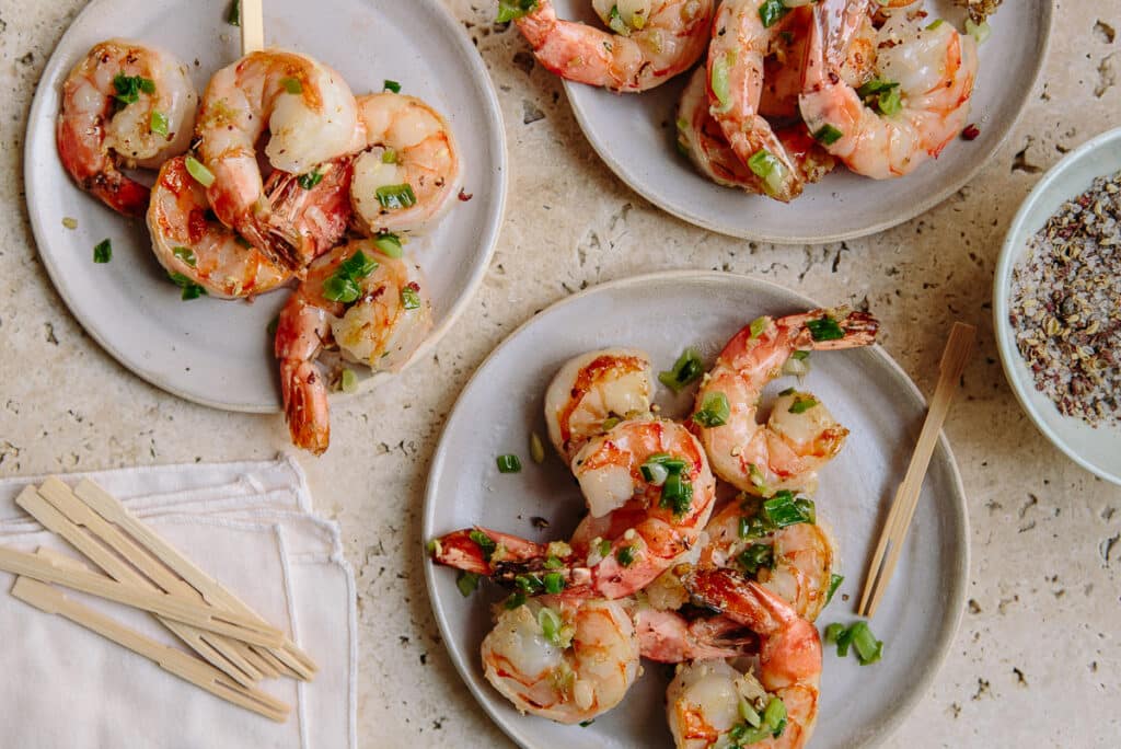 Grilled shrimp garnished with chopped green onions served on beige plates, accompanied by wooden skewers and a bowl of seasoning, showcasing a delicious seafood dish perfect for appetizers or gatherings.
