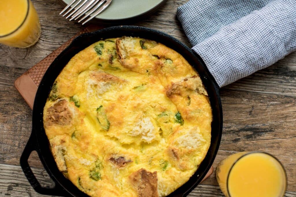 Delicious baked egg frittata with bread and greens in a cast iron skillet, served with glasses of orange juice and a textured napkin, perfect for a brunch setting.