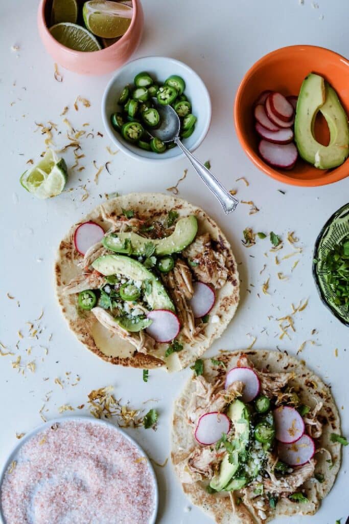 Two tacos filled with shredded chicken, avocado slices, radishes, jalapeños, and cilantro on a white surface. Accompanying ingredients include lime wedges, a bowl of sliced jalapeños, and a dish of pink salt, creating a vibrant and appetizing scene.
