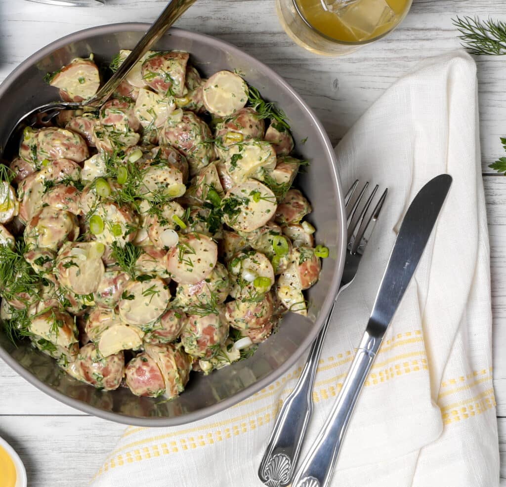Creamy potato salad with fresh herbs, garnished with green onions and dill, served in a large bowl on a wooden table, accompanied by utensils and a refreshing drink.