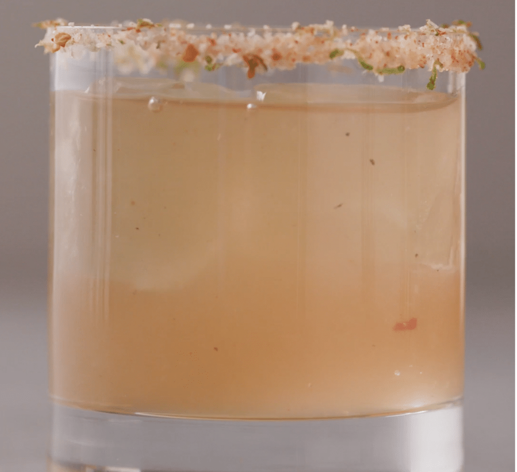 Close-up of a refreshing cocktail in a clear glass, featuring a salted rim and ice cubes, with a light amber liquid that hints at citrus flavors and garnished with herbs.