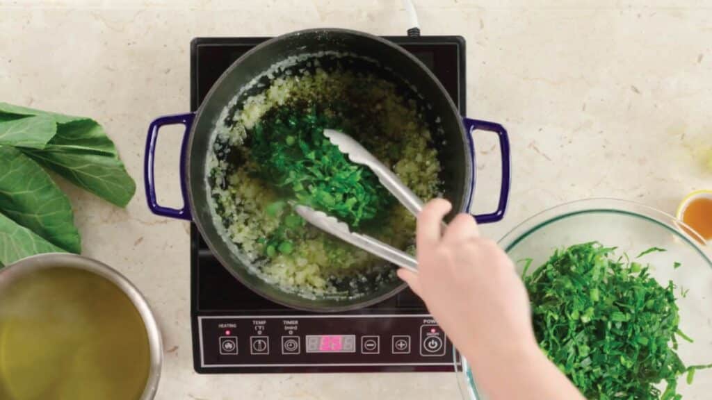 Cooking greens in a pot on an induction stove, with a hand using tongs to mix chopped leafy vegetables, surrounded by fresh greens and broth.