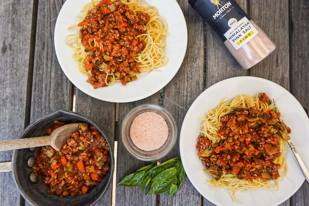 Two plates of spaghetti topped with a rich meat sauce made from ground beef, carrots, and bell peppers, served on a rustic wooden table. A pot of sauce with a wooden spoon, a bowl of pink Himalayan salt, and fresh basil leaves are also visible, enhancing the meal's presentation.