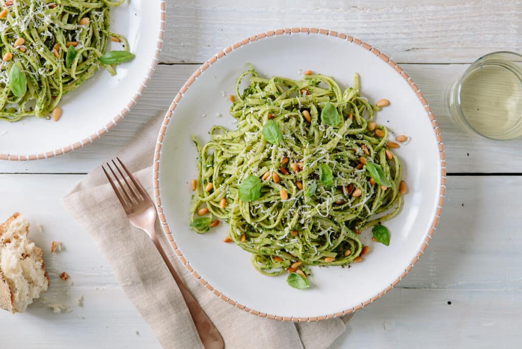 Delicious plate of spaghetti with green pesto, garnished with fresh basil leaves and pine nuts, served on a rustic wooden table alongside a glass of white wine and a piece of bread.