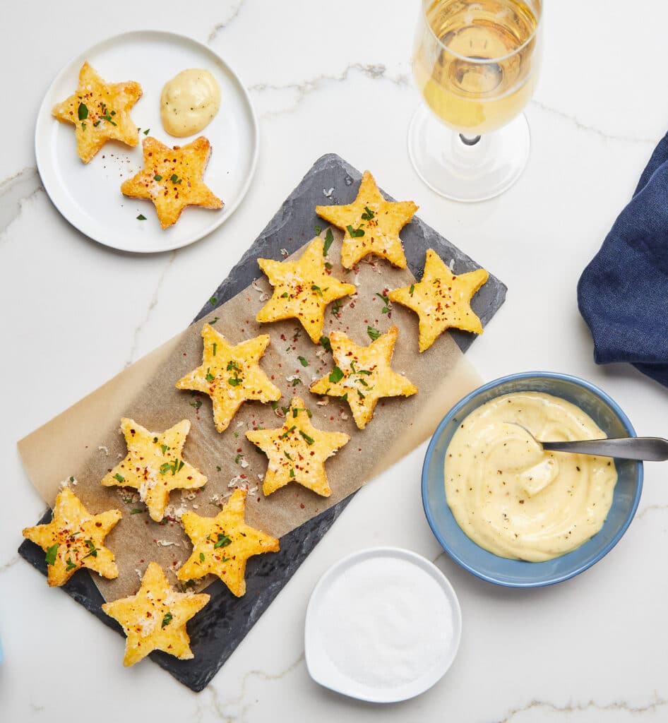 Star-shaped crispy snacks arranged on a slate platter, garnished with herbs and red pepper flakes, served with a creamy dipping sauce and a side of salt. A glass of white wine and additional star-shaped snacks on a white plate complete the table setting. Ideal for appetizers or party food.
