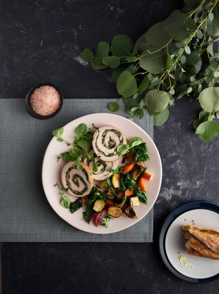 A beautifully arranged plate featuring sliced herb-stuffed chicken, accompanied by a colorful medley of roasted vegetables, including carrots, Brussels sprouts, and kale, garnished with fresh greens. A small bowl of pink salt and a side of toasted bread complete the presentation.