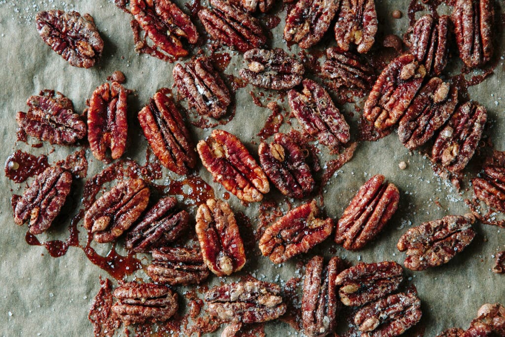 Roasted pecans coated in a sweet glaze, sprinkled with sea salt, arranged on parchment paper. Ideal for snacks, desserts, or salad toppings, showcasing a rich brown color and glossy finish.