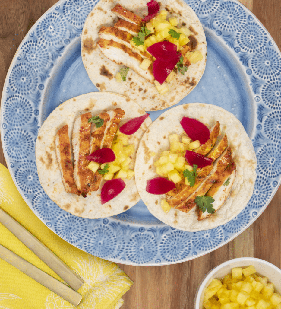 Three delicious chicken tacos on a blue decorative plate, topped with fresh diced pineapple, pickled onions, and cilantro. A small bowl of additional pineapple sits nearby, accompanied by gold utensils and a yellow napkin, creating an inviting meal setting.