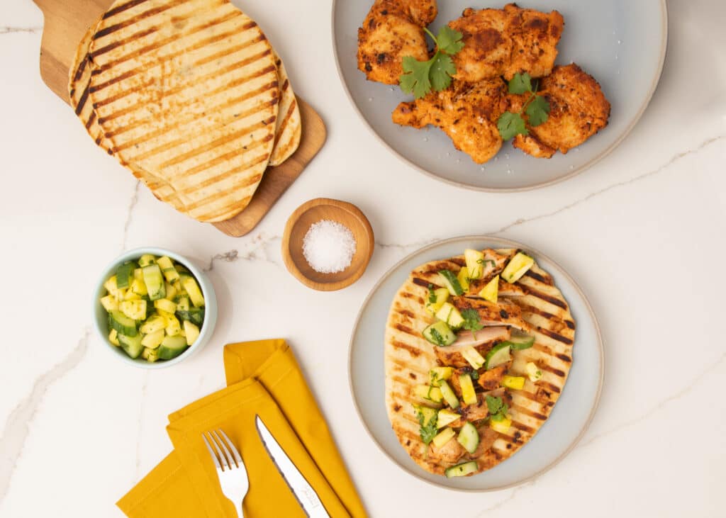 Grilled flatbreads served with spiced chicken, fresh cucumber, and pineapple salsa on a marble countertop, accompanied by salt and yellow napkins.