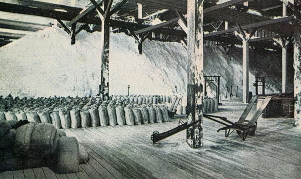 Interior view of a salt storage facility featuring large piles of salt and neatly arranged burlap sacks. The structure has wooden beams and a wooden floor, with various tools and equipment visible, indicating a working environment focused on salt production and storage.