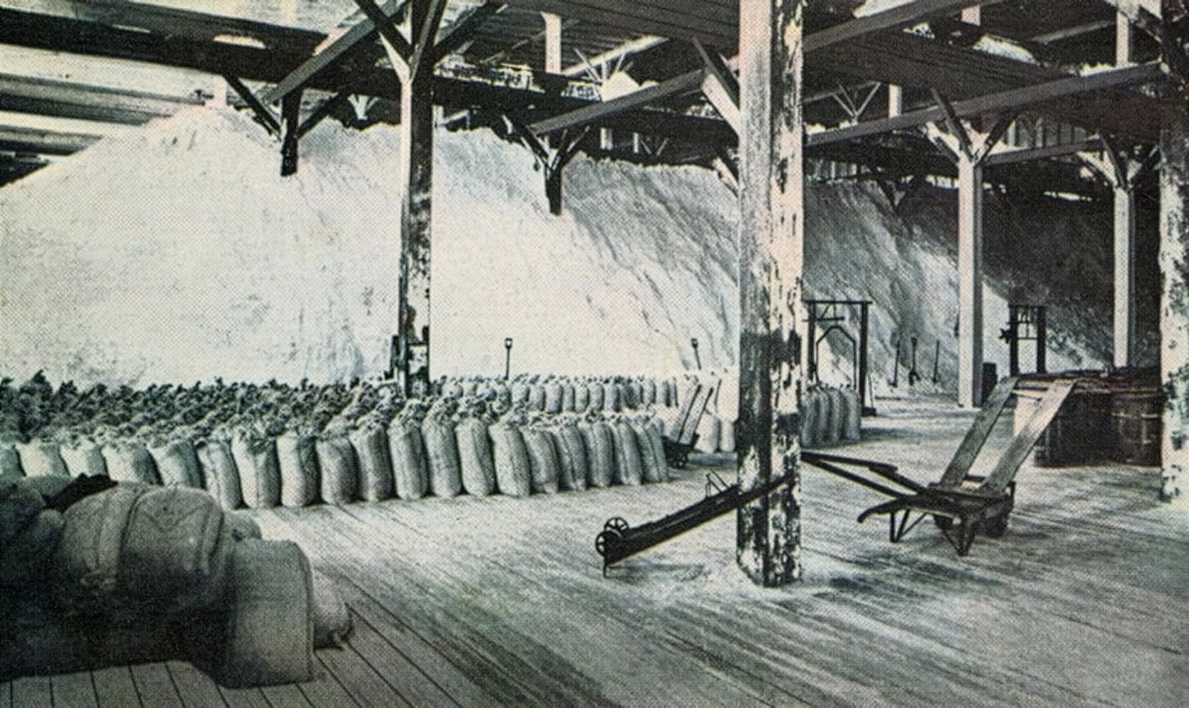Interior view of a salt storage facility featuring large piles of salt and neatly arranged burlap sacks. The structure has wooden beams and a wooden floor, with various tools and equipment visible, indicating a working environment focused on salt production and storage.