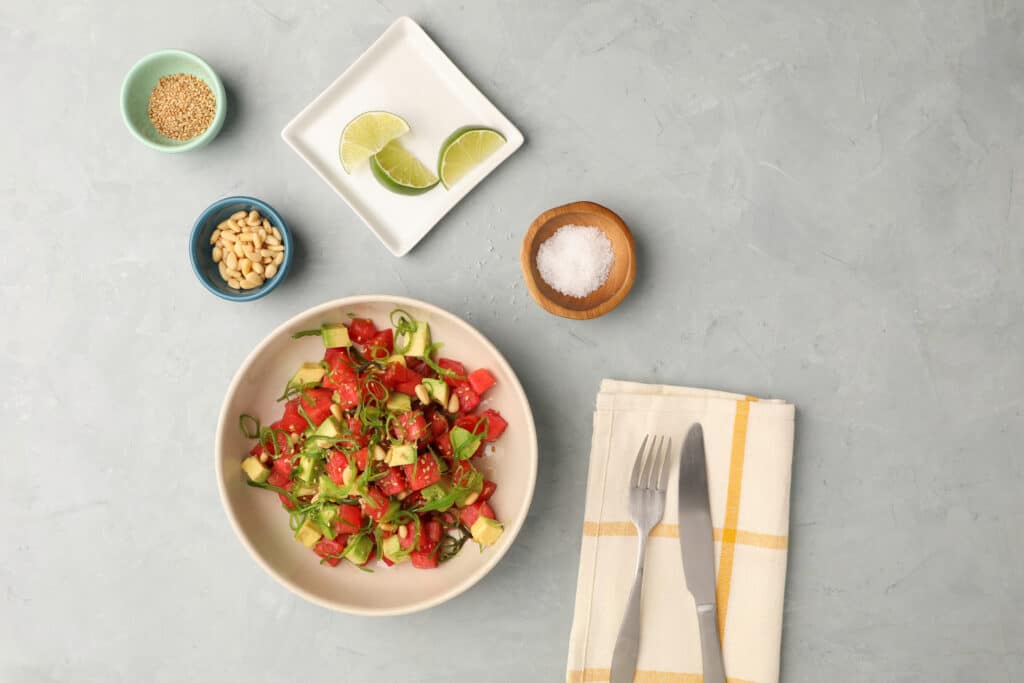 Fresh vegetable salad in a bowl, featuring diced tomatoes, cucumber, and green onions, topped with pine nuts and sesame seeds. Accompanied by lime wedges, salt in a wooden bowl, and utensils on a checkered napkin, all arranged on a textured gray surface.