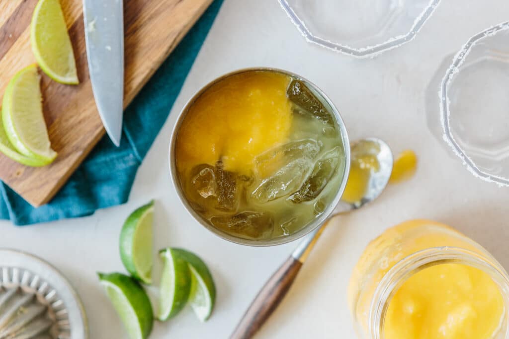A refreshing cocktail with mango puree and ice, served in a metal glass, surrounded by fresh lime wedges, a knife, and a jar of mango puree on a light countertop.