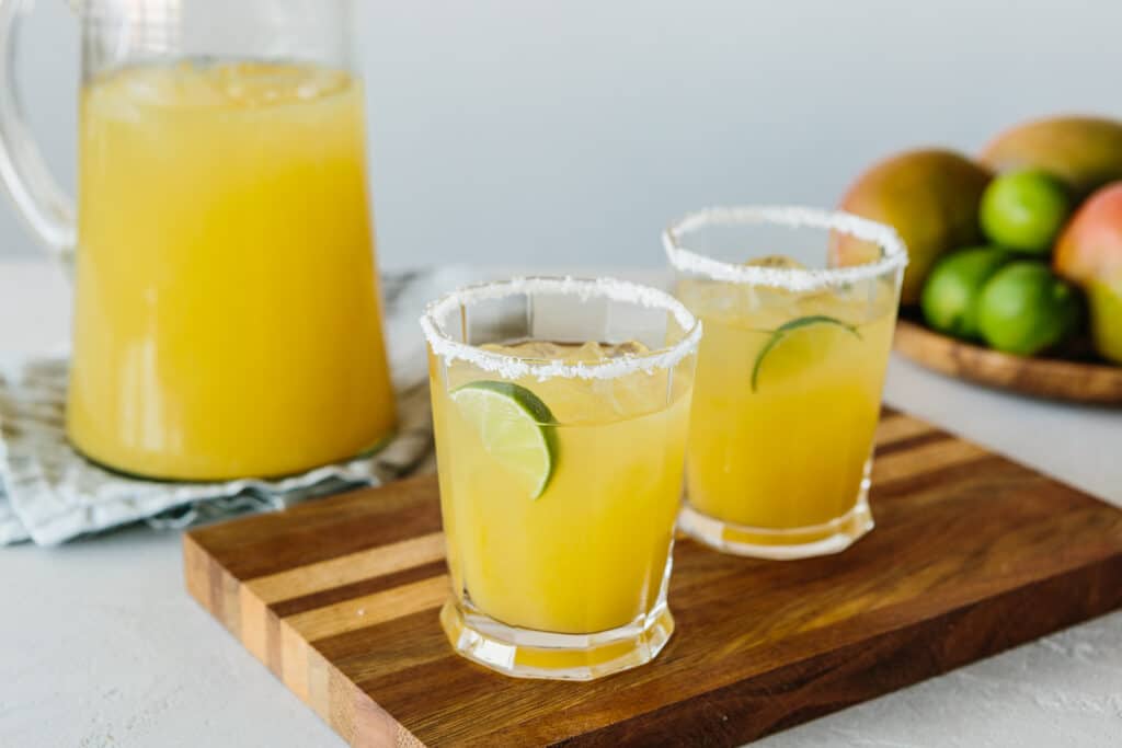 Refreshing mango margaritas served in glasses with salted rims, garnished with lime slices, alongside a pitcher of mango juice and a bowl of fresh fruits on a wooden cutting board.