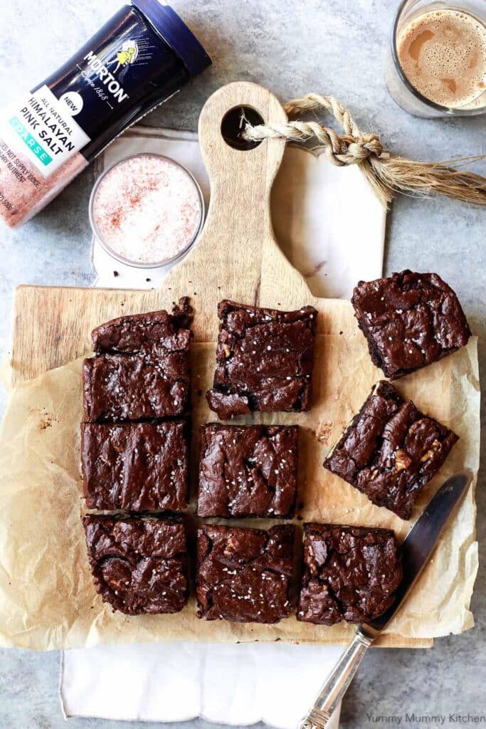 Delicious homemade brownies cut into squares, topped with flaky sea salt, displayed on a wooden cutting board alongside a glass of pink Himalayan salt and a cup of coffee. Perfect for dessert lovers and baking enthusiasts.