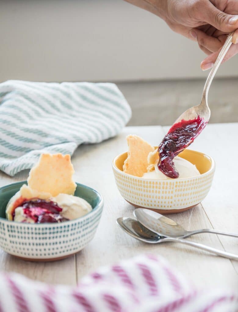 A hand scooping berry jam onto vanilla ice cream in a colorful bowl, with another bowl of ice cream and cookies in the background, on a wooden table with a striped cloth and silver spoons.