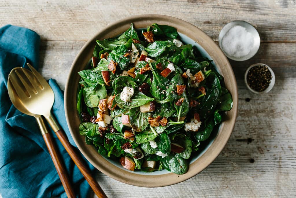 Fresh spinach salad topped with crispy bacon, diced apples, and crumbled feta cheese, served in a rustic bowl with a side of salt and pepper. Golden serving utensils and a blue napkin accompany the dish on a wooden table.