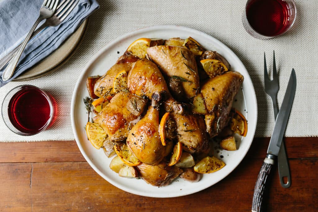 Roasted chicken legs with lemon and herbs served on a white plate, accompanied by golden potatoes and garnished with fresh rosemary. A glass of red wine and silverware are set on a rustic wooden table, creating a cozy dining atmosphere.