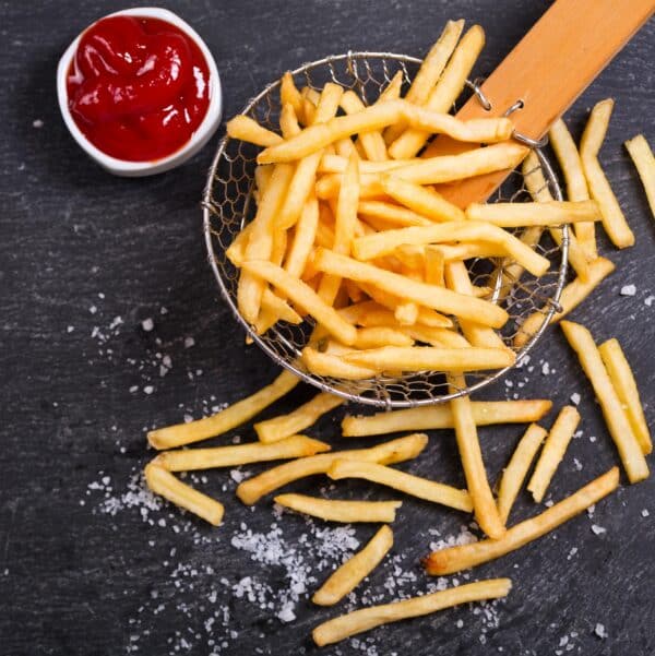 Crispy golden French fries in a wire basket with a wooden handle, surrounded by scattered fries and a small bowl of ketchup on a dark slate background.