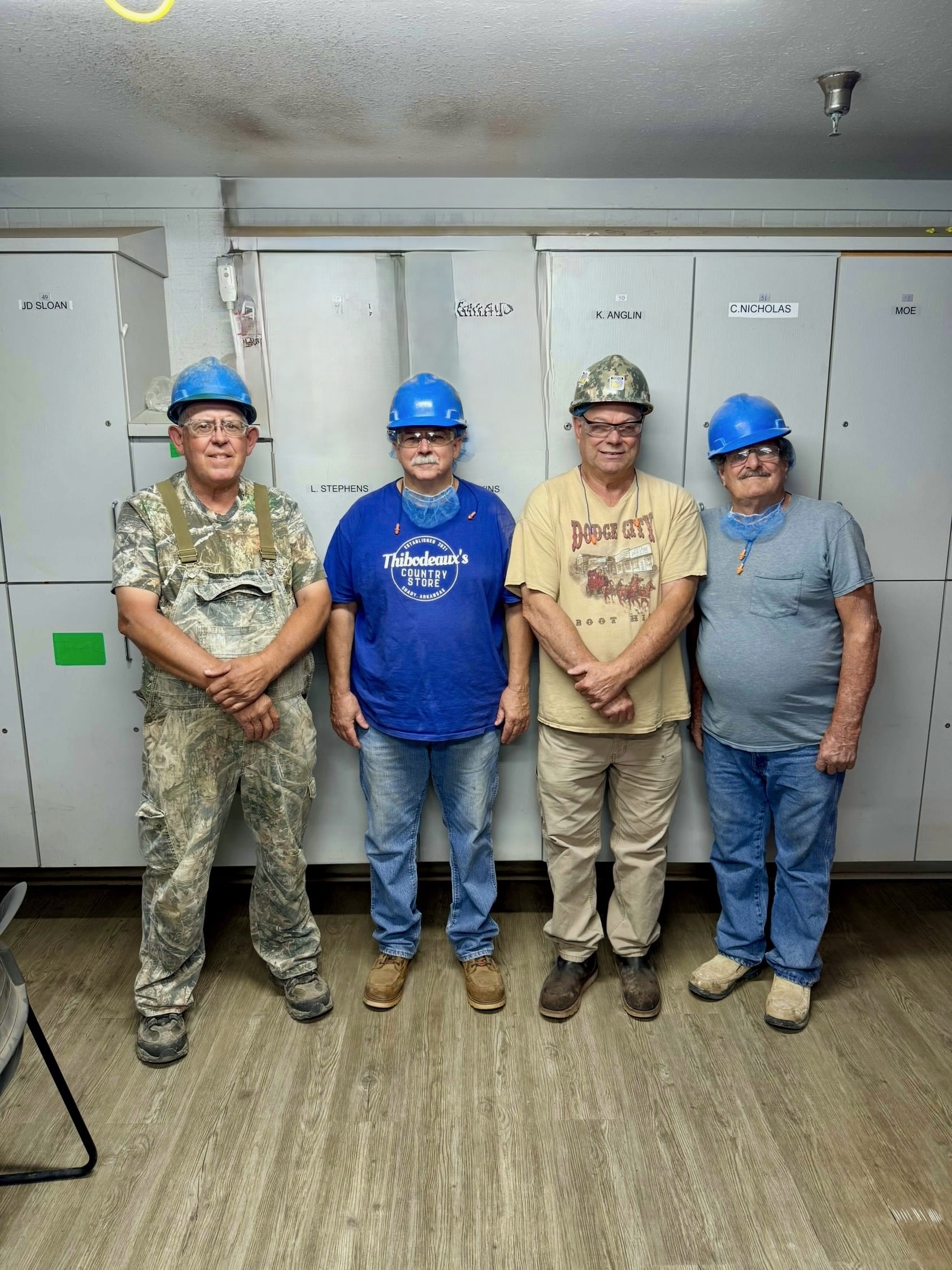 Quatre hommes casqués, vêtus de tenues de travail, posent en intérieur, avec des armoires de rangement en arrière-plan. Le lieu semble être un lieu de travail, probablement lié à la construction ou aux mines.