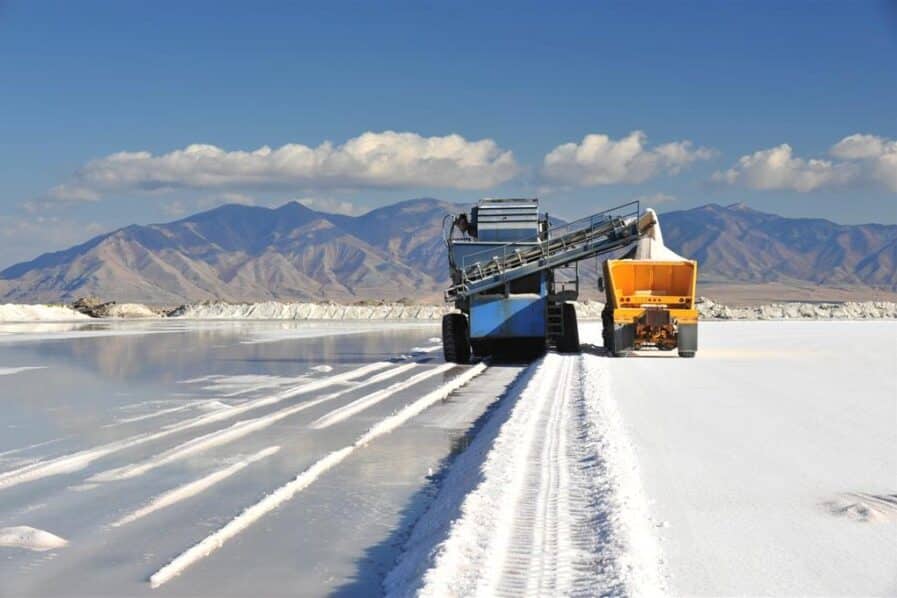 Salt harvesting equipment working on a salt flat with mountains in the background, showcasing a blue and yellow vehicle transporting salt along a reflective surface.
