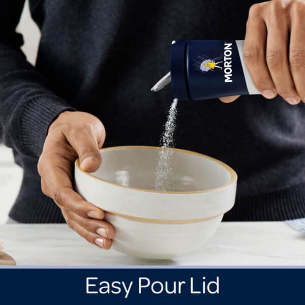 Person pouring salt from a Morton container with an easy pour lid into a ceramic bowl on a kitchen countertop.