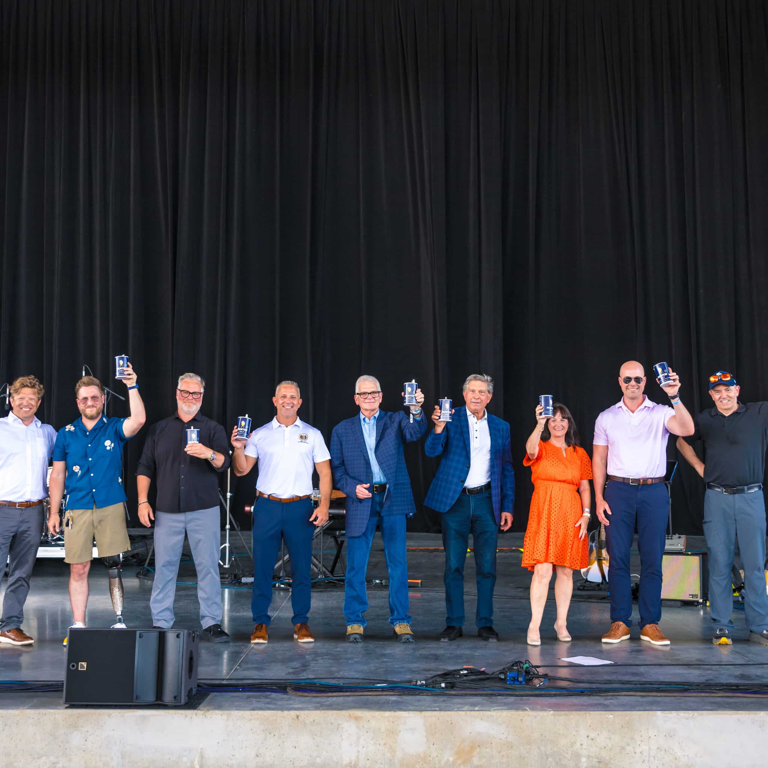 Group of individuals standing on stage, holding up drinks in celebration. The backdrop features a black curtain, with musical instruments visible on the side. The event appears festive and community-oriented.