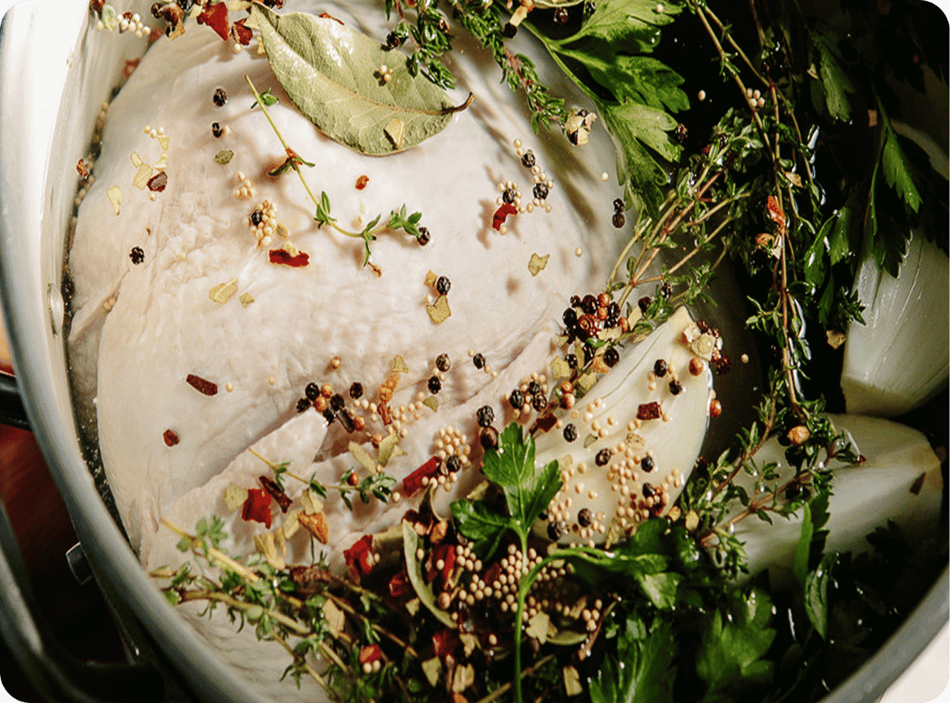 Raw chicken submerged in a pot with fresh herbs and spices, including bay leaves, thyme, and parsley, ready for cooking.