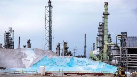 Raffinerie industrielle avec d'immenses tas de produits chimiques aux reflets bleutés et d'imposants équipements de traitement sous un ciel nuageux.