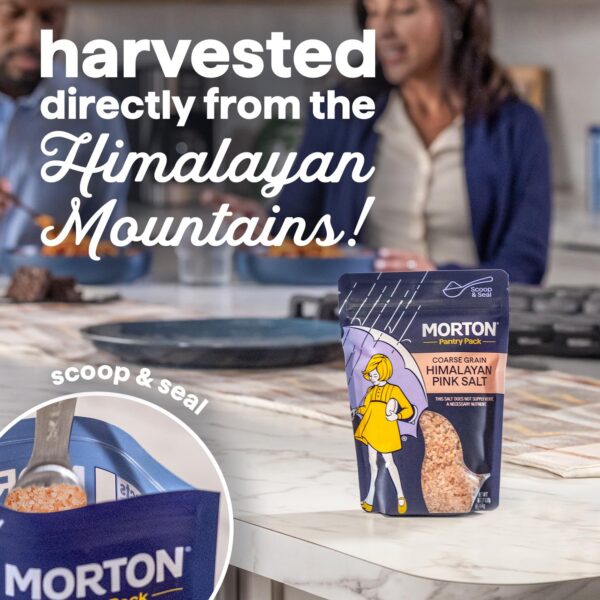 Morton Himalayan Pink Salt packaging on a kitchen countertop, showcasing coarse grain salt harvested from the Himalayan Mountains. The image features a spoon scooping salt from the package, emphasizing the "scoop & seal" design. In the background, two people enjoy a meal, highlighting the salt's culinary use.