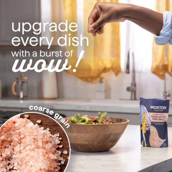 Hand sprinkling coarse grain Himalayan pink salt over a fresh salad in a wooden bowl, with a Morton salt package displayed prominently on a kitchen countertop. The image emphasizes enhancing dishes with flavorful seasoning.