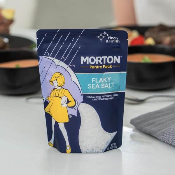 Morton Flaky Sea Salt Pantry Pack displayed on a kitchen countertop, with a blurred background of bowls containing food. The packaging features a playful design with a character holding an egg, emphasizing its culinary use. Ideal for enhancing flavors in various dishes.