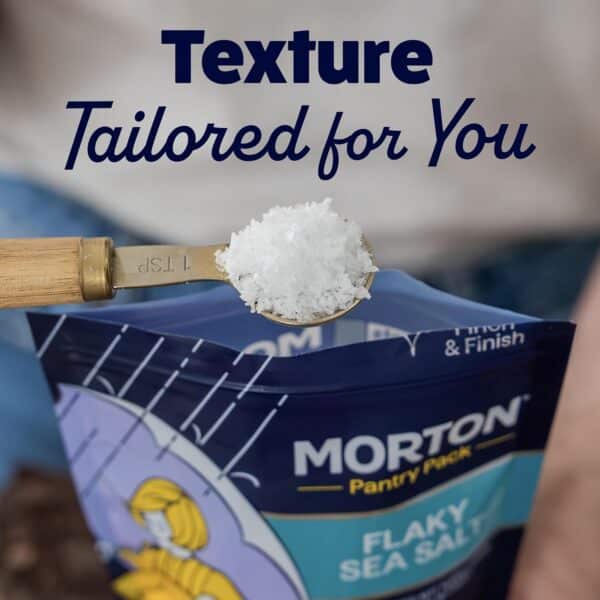 Spoonful of flaky sea salt above a Morton Pantry Pack, showcasing the product's texture and packaging design for culinary use.