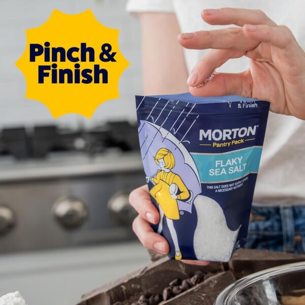 Person holding a bag of Morton Flaky Sea Salt in a kitchen setting, demonstrating the "Pinch & Finish" technique for enhancing dishes. The background features cooking equipment, emphasizing culinary use.