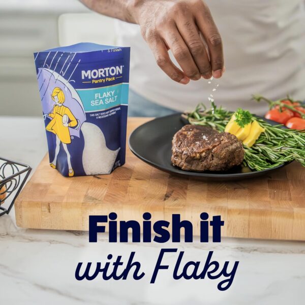 A hand sprinkles Morton Flaky Sea Salt over a perfectly cooked steak on a black plate, surrounded by fresh rosemary and cherry tomatoes, highlighting the seasoning's role in enhancing flavor.