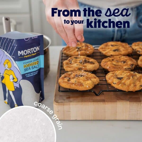 Hand sprinkling Morton coarse grain iodized sea salt over freshly baked chocolate chip cookies cooling on a wire rack, with a package of salt prominently displayed in the background.