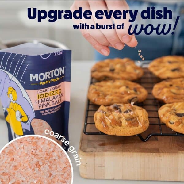 Hand sprinkling Morton Coarse Grain Iodized Himalayan Pink Salt over freshly baked cookies cooling on a wire rack, showcasing the salt packaging and highlighting its ability to enhance flavors in dishes.