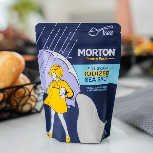 Morton Pantry Pack fine grain iodized sea salt in a colorful packaging featuring a girl with an umbrella, designed for easy scooping and sealing. Ideal for enhancing flavor and supplying essential iodine, set against a backdrop of freshly baked bread.
