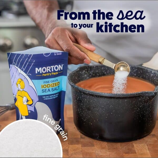 Hand pouring Morton fine grain iodized sea salt from a measuring spoon into a pot, showcasing the product's packaging and emphasizing its journey from the sea to the kitchen.
