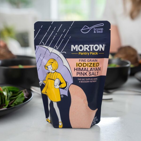 Morton Pantry Pack fine grain iodized Himalayan pink salt in a colorful pouch, featuring a playful illustration of a girl with an umbrella, set against a kitchen background with food items. Ideal for enhancing flavor and providing essential iodine in cooking.