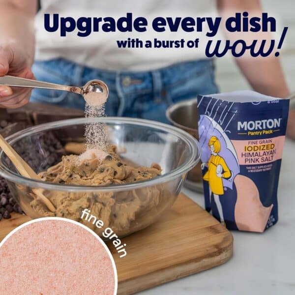 Person adding fine grain iodized Himalayan pink salt to cookie dough in a glass bowl, with a package of Morton salt in the background, emphasizing the flavor enhancement in cooking.