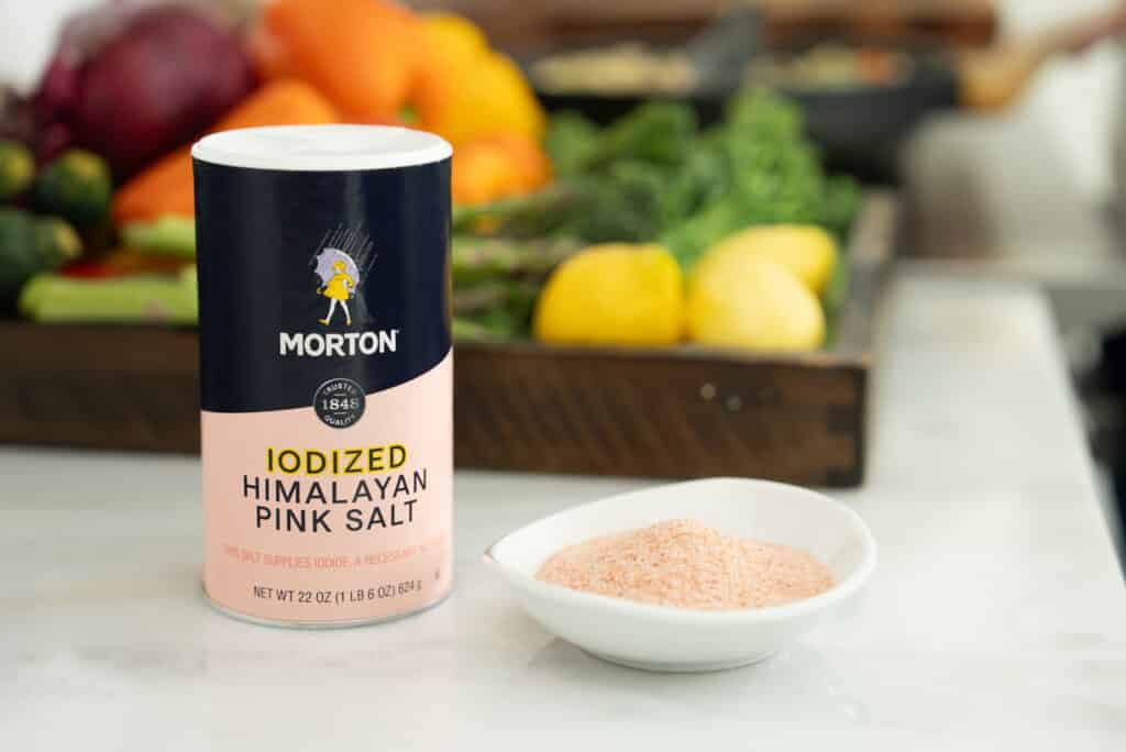 Morton Iodized Himalayan Pink Salt container next to a bowl of pink salt, with fresh vegetables and fruits in the background, highlighting the salt's culinary and nutritional benefits.
