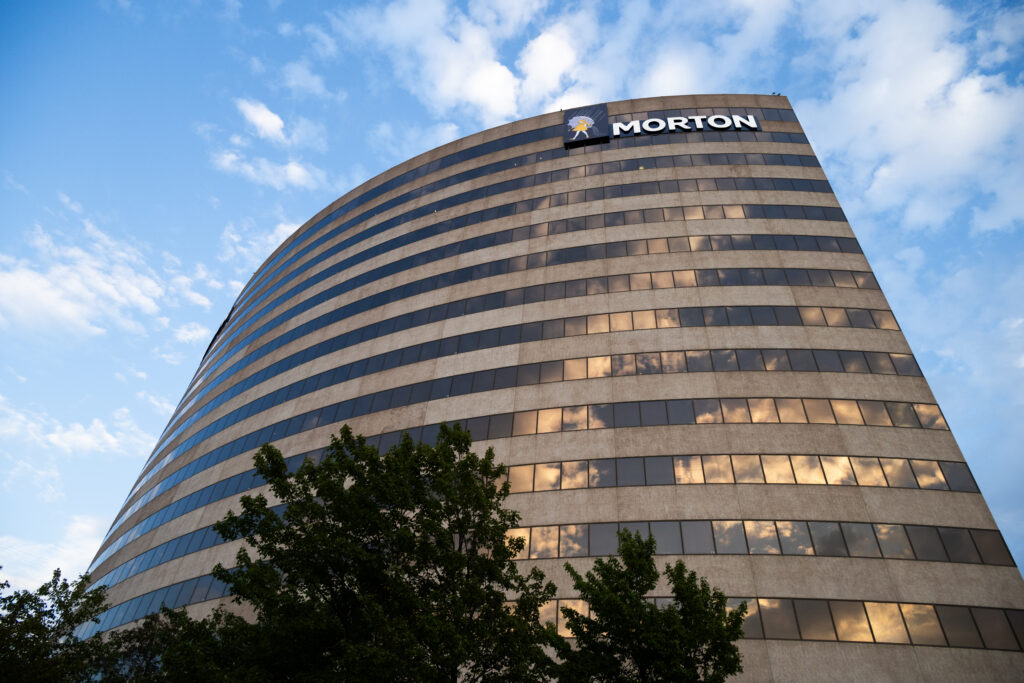 Modern office building with a curved facade featuring the "Morton" logo, surrounded by greenery and reflecting the sky in its windows.