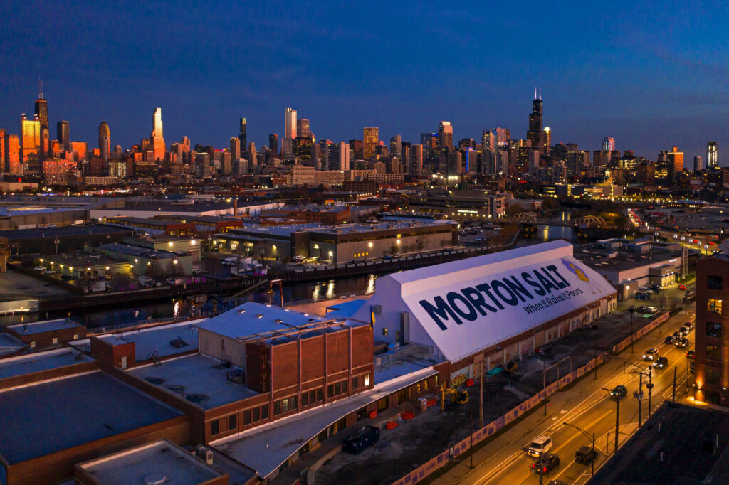 Aerial view of the Morton Salt facility at dusk, featuring the iconic building with a prominent sign, surrounded by the Chicago skyline illuminated by city lights. The image captures the urban landscape with streets and nearby structures visible.