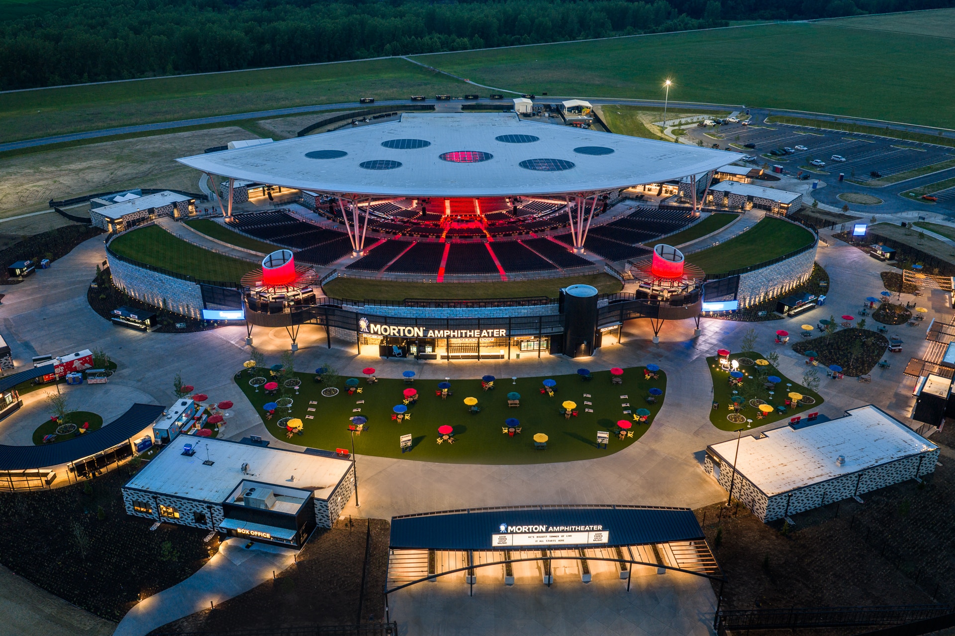Aerial view of the Morton Amphitheater showcasing its modern design, outdoor seating areas with colorful umbrellas, and a spacious parking lot, surrounded by lush greenery. The amphitheater features a unique roof structure and vibrant lighting, creating an inviting atmosphere for concerts and events.
