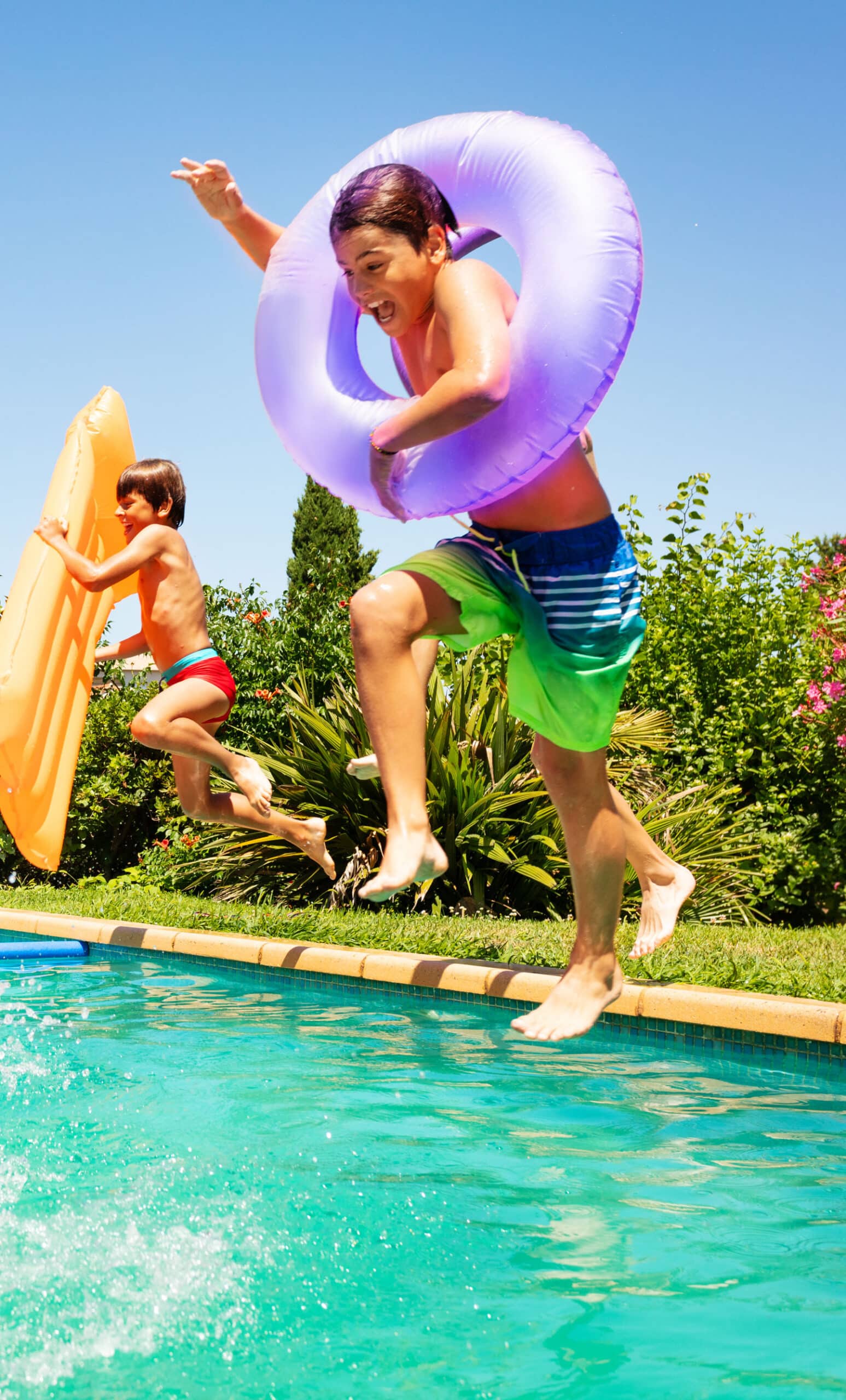 Two children joyfully jumping into a swimming pool on a sunny day, one wearing a purple inflatable ring and the other holding an orange float. Lush greenery and colorful flowers surround the pool area, creating a vibrant summer atmosphere.
