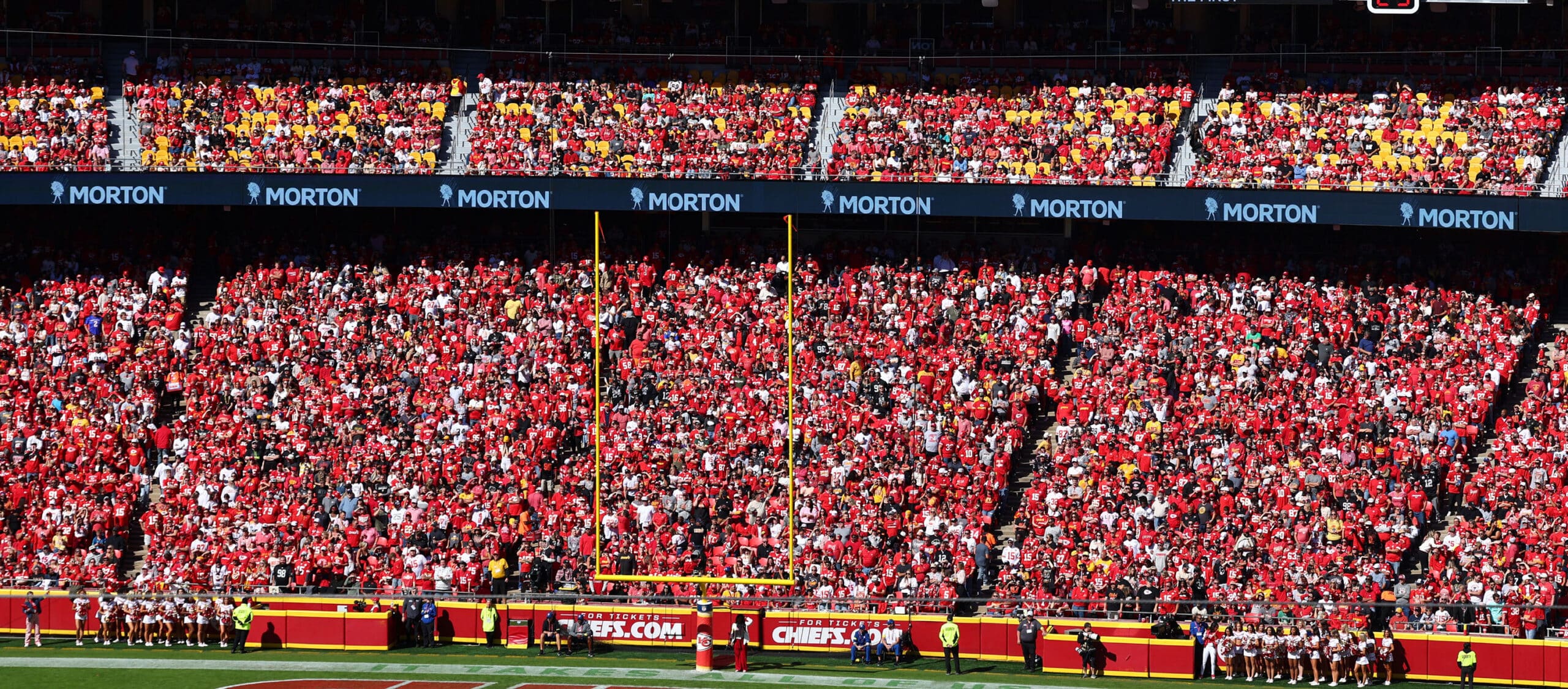 A vibrant crowd of fans dressed in red fills the stadium, showcasing team spirit during a football game. The stands are packed with supporters, while cheerleaders and security personnel are visible near the field. Bright banners and goalposts are prominently displayed, emphasizing the energetic atmosphere of the event.