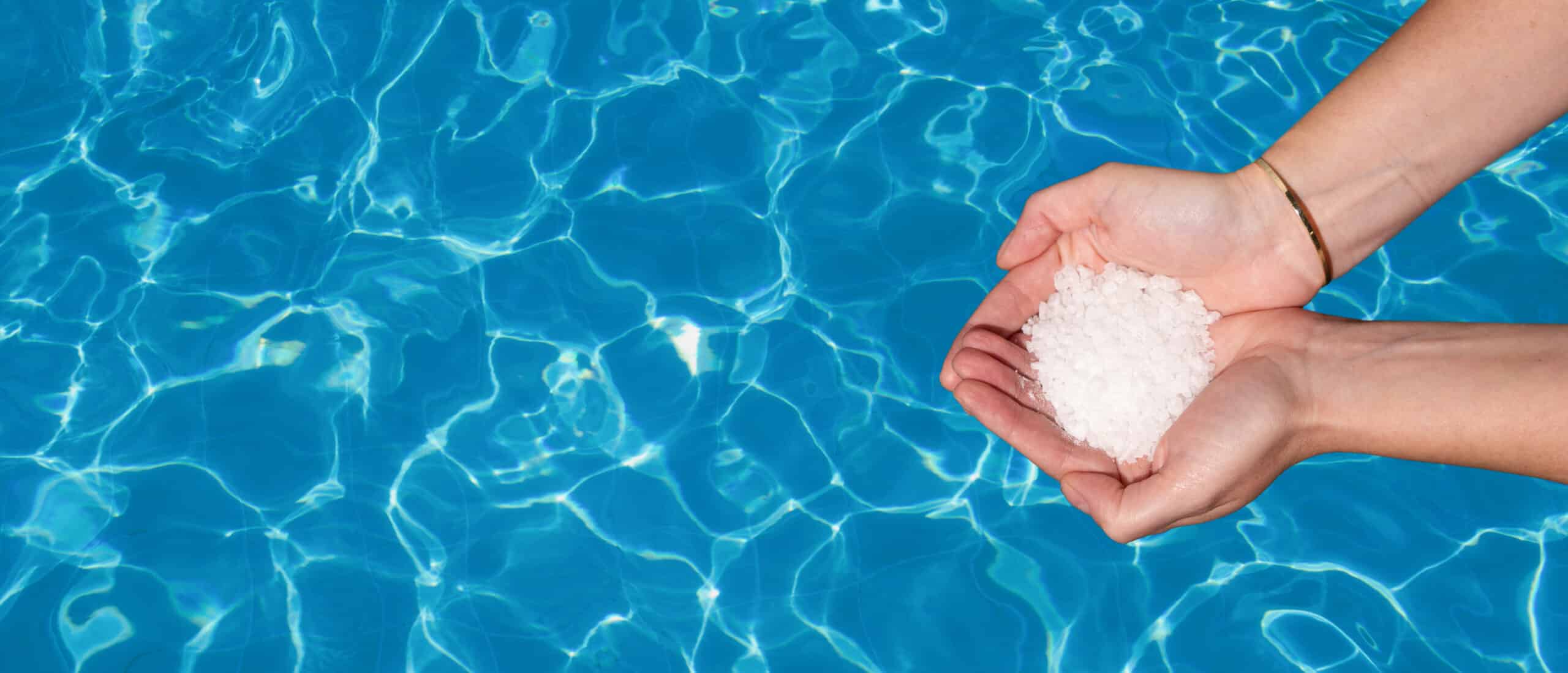 Hands holding pool salt above a sparkling blue swimming pool, emphasizing the relaxation and wellness benefits of salt in water.
