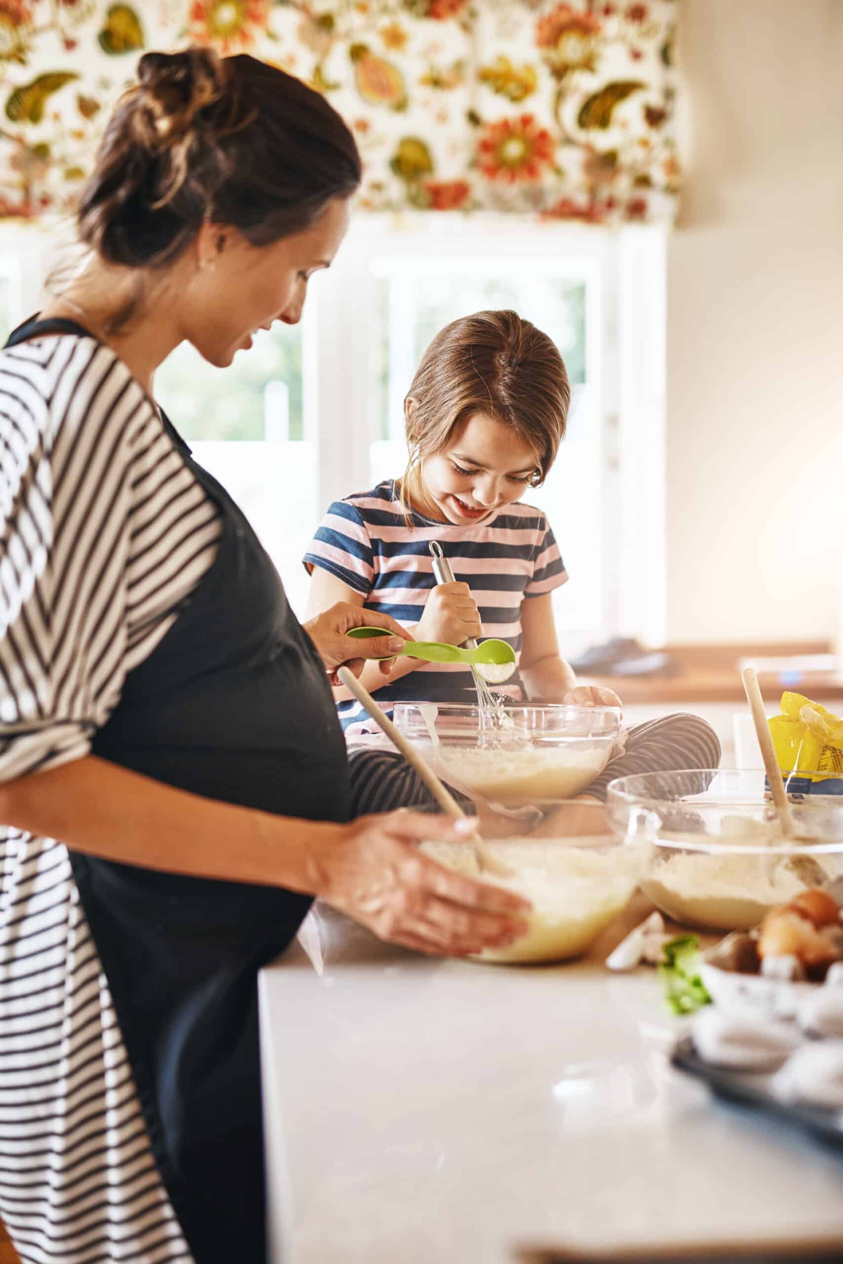 A pregnant woman and a young girl are joyfully baking together in a bright kitchen, mixing ingredients in large bowls. The girl is using a whisk, while the woman stirs with a spoon, showcasing a warm family bonding moment through cooking.