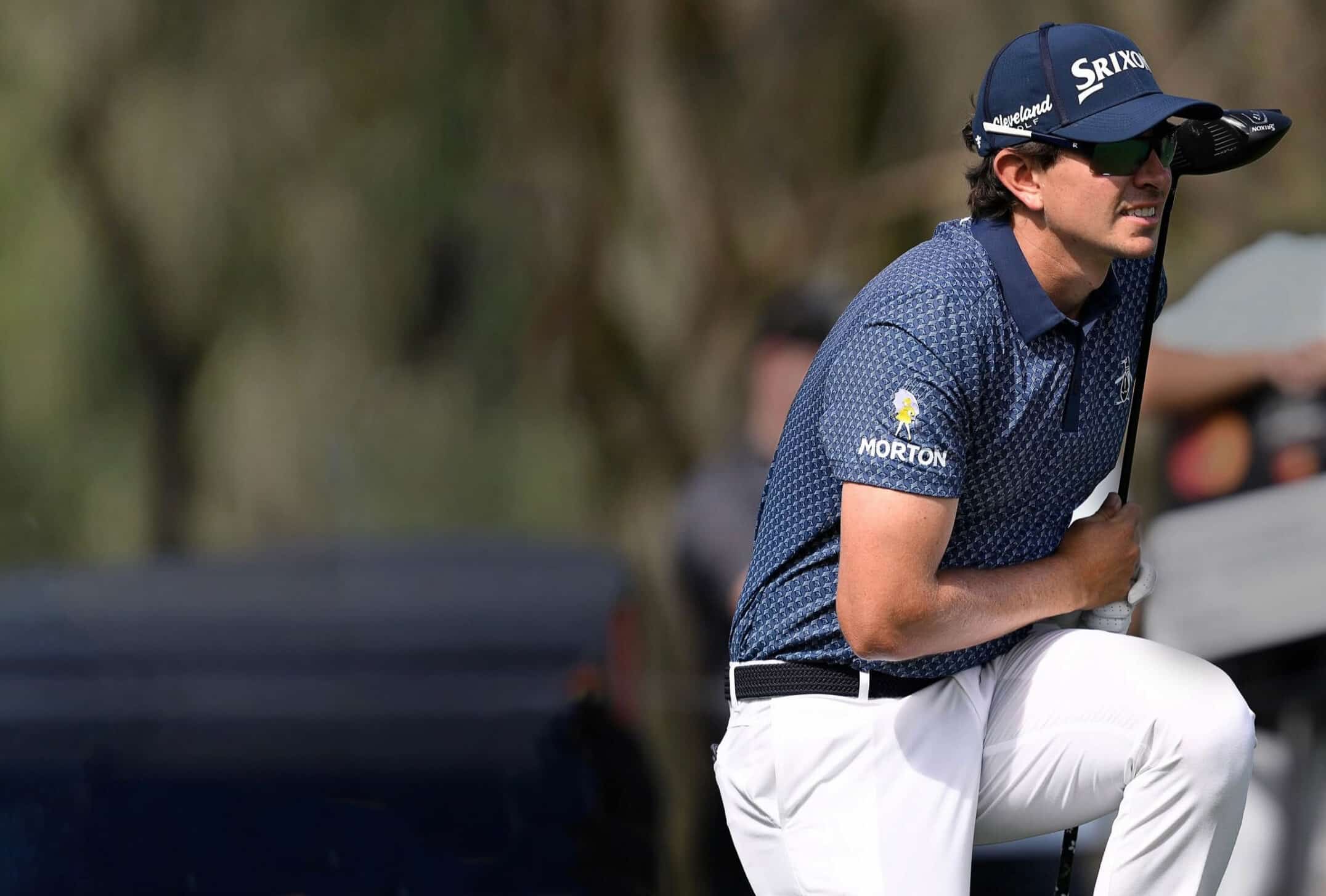 Professional golfer preparing to take a shot, wearing a blue patterned shirt and white pants, with a golf club in hand. The background features blurred trees, indicating an outdoor golf course setting.